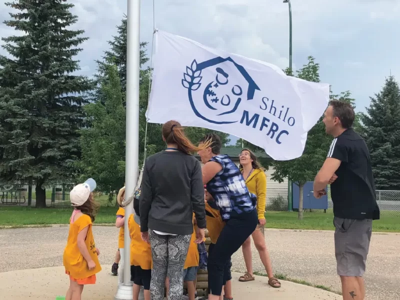 Custom Flag 02 | The Flag Shop Photo of children and teachers raising a white flag with blue text "Shilo MFRC" on a flagpole
