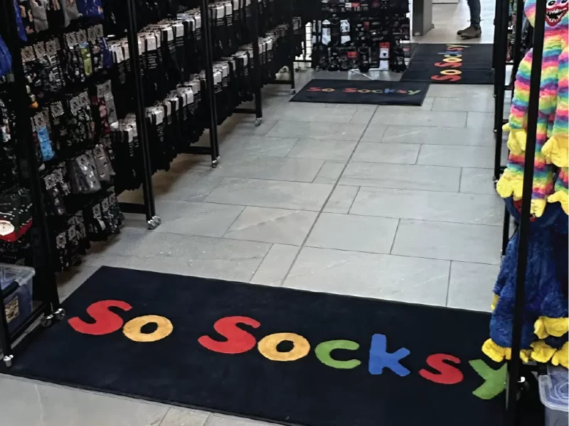 Custom Floor Mats | The Flag Shop Custom floor mat on display at a retail location. Black mat with multi coloured "So Socksy" logo